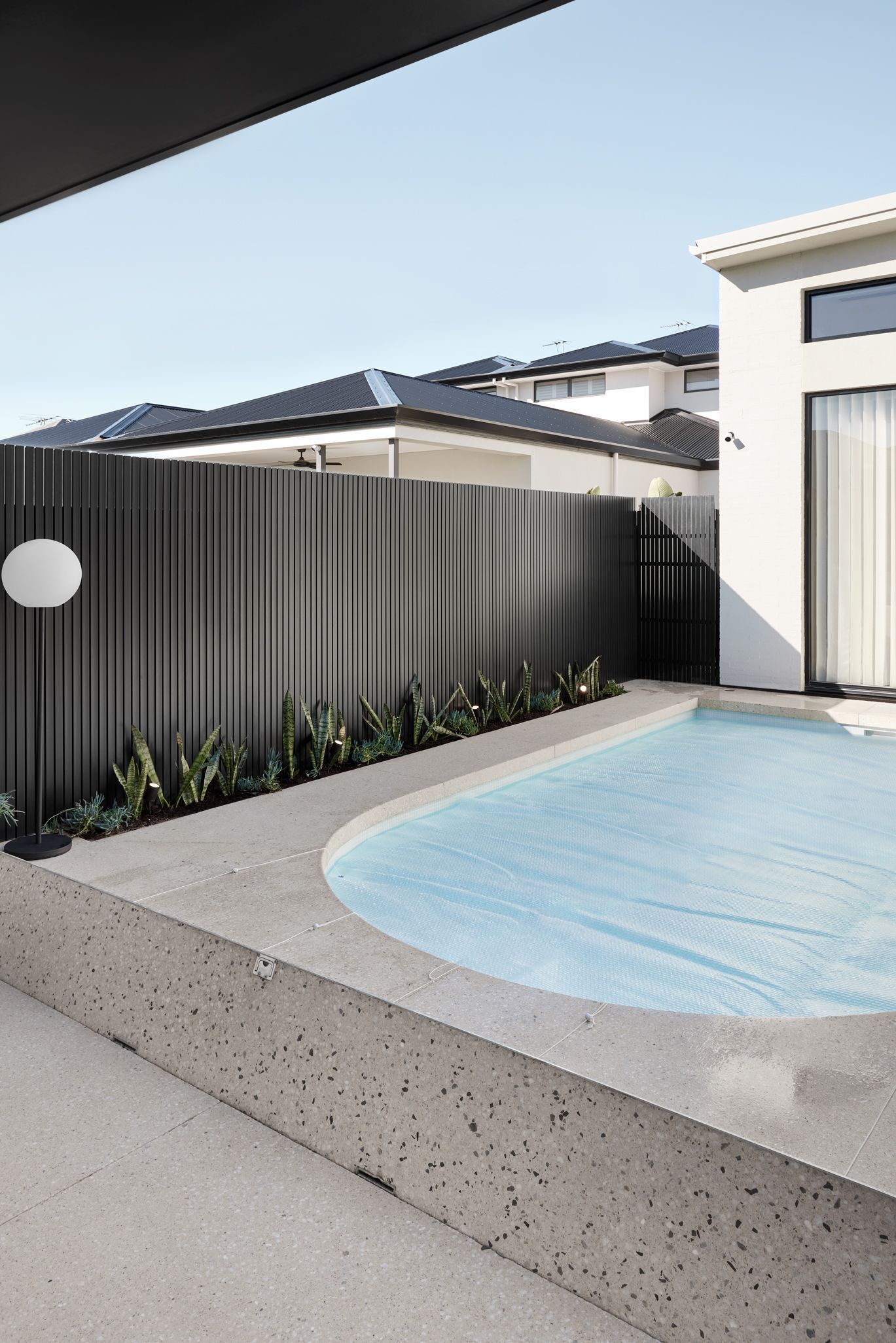 Pool area with a concrete edge, dark fence, and a white house in the background on a sunny day.
