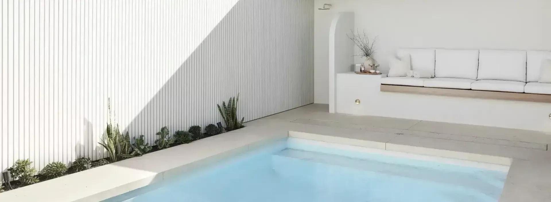 A minimalist, all-white outdoor pool area with a small pool, bench with pillows, and slatted wall.