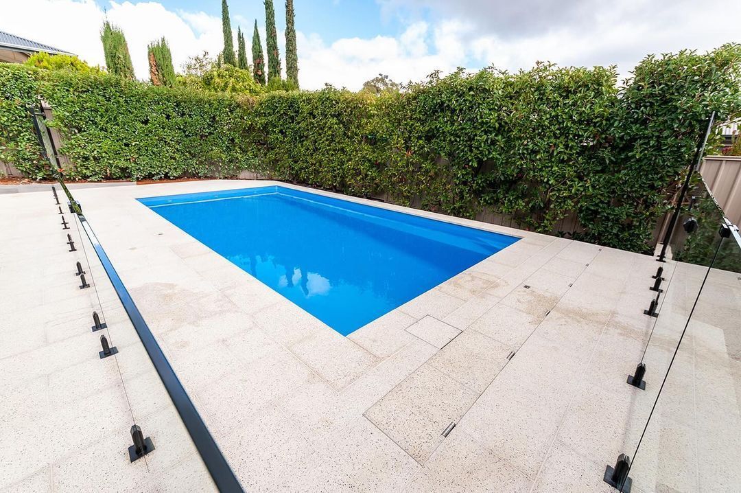Rectangular blue swimming pool surrounded by stone tiles and a green hedge under a bright sky.
