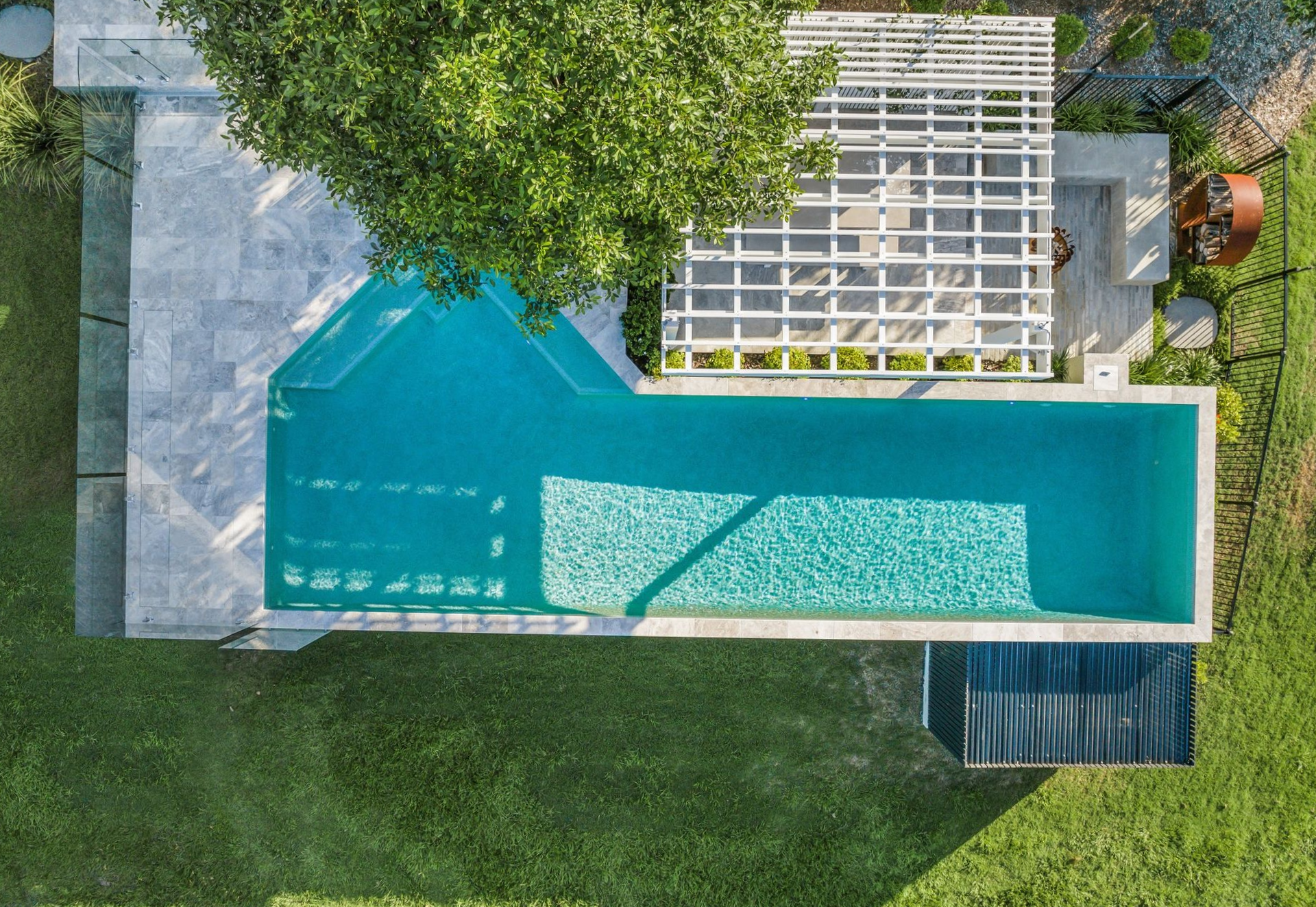 Overhead view of a rectangular pool with turquoise water, a white pergola, and green grass.