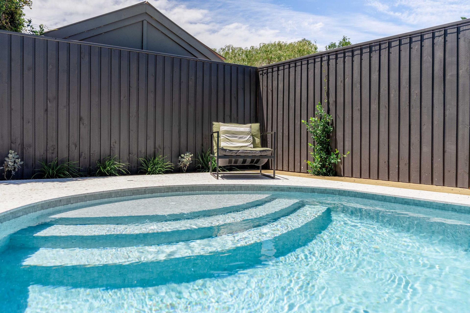 Pool with steps, light blue water, and a wooden chair against a dark fence.
