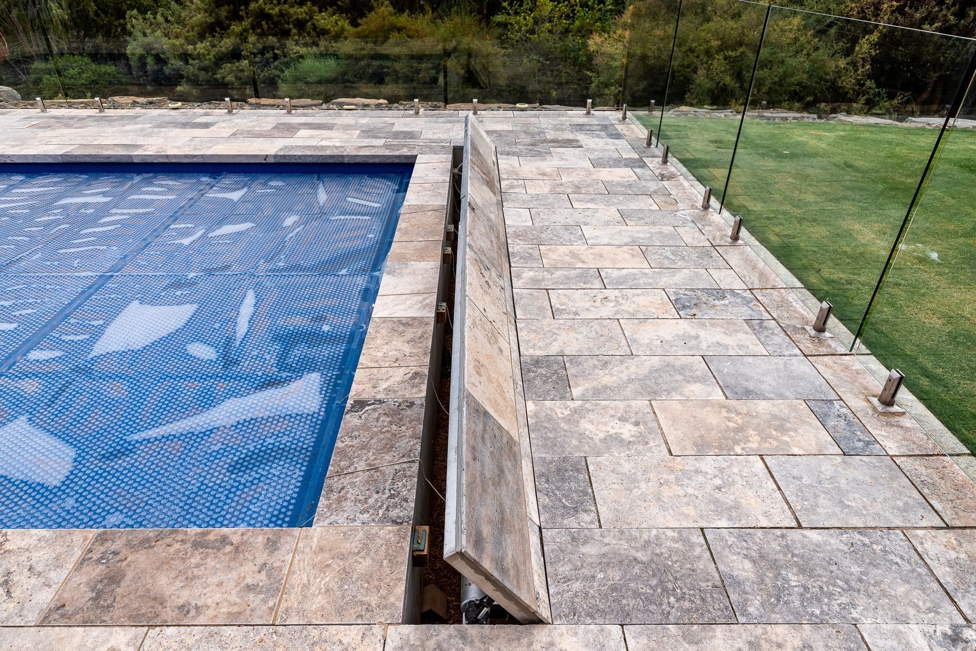 Pool with blue cover, stone patio, and glass fence along a grassy yard.