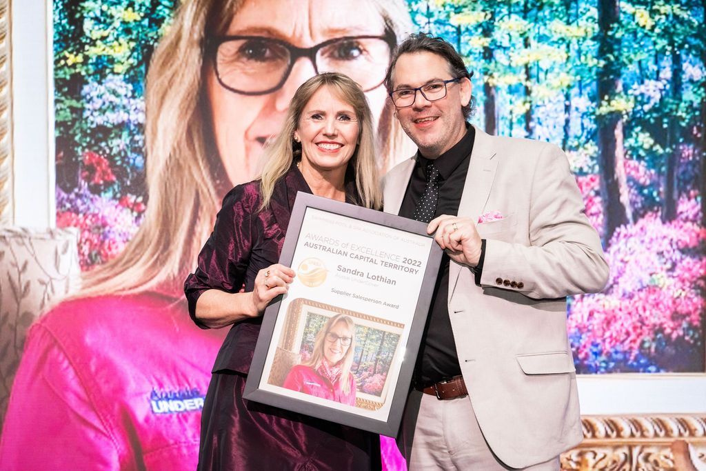 Woman and man hold award, smiling, against a backdrop with a woman's image.