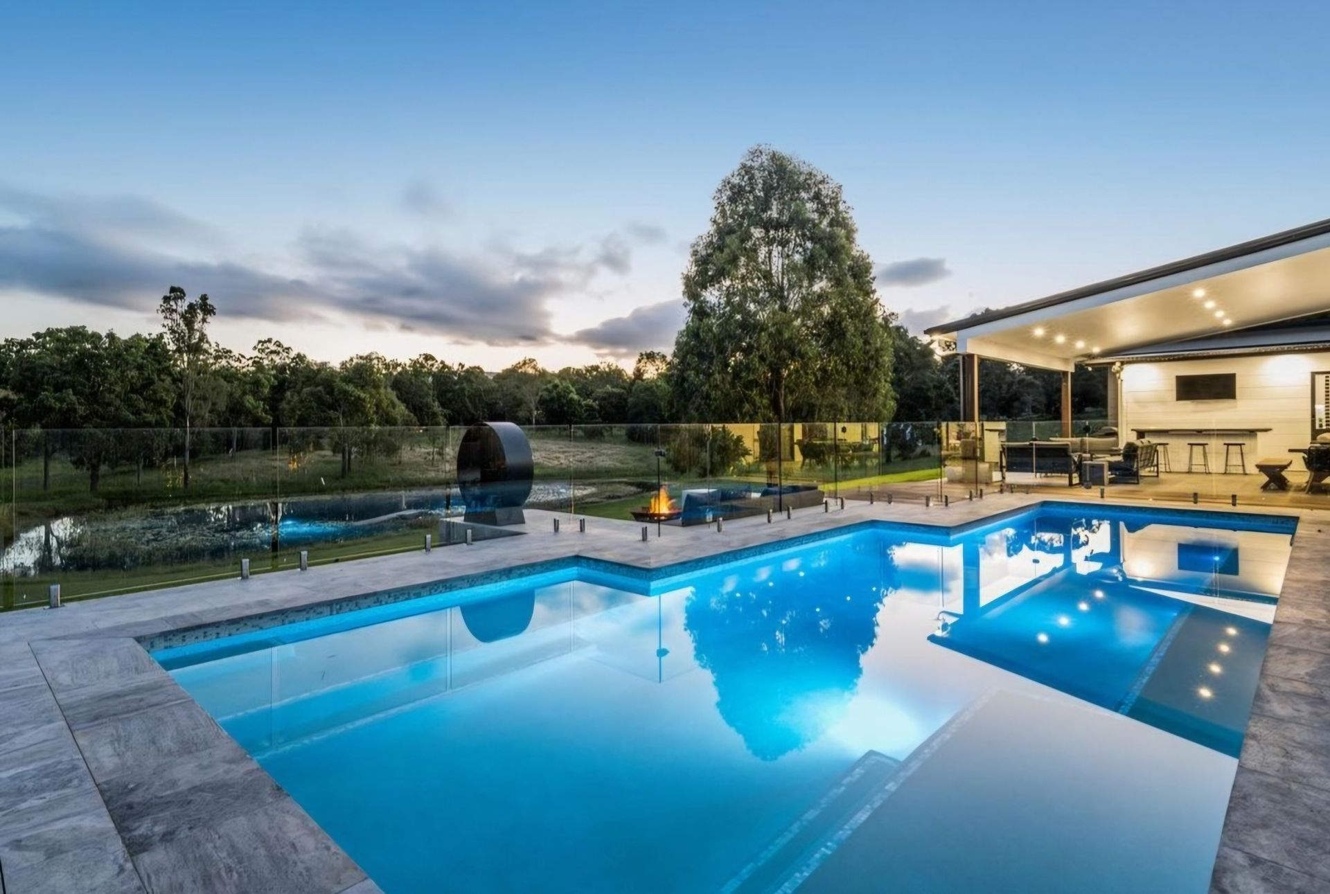 Pool with blue underwater lights, near a home at dusk with trees in the background.