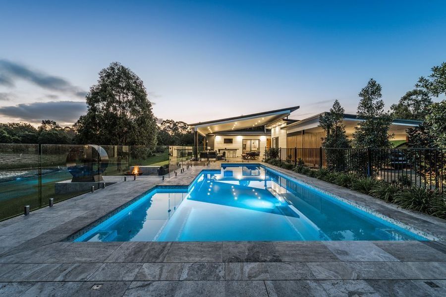 A modern home with a rectangular pool, nighttime, blue pool lights, and a lawn.