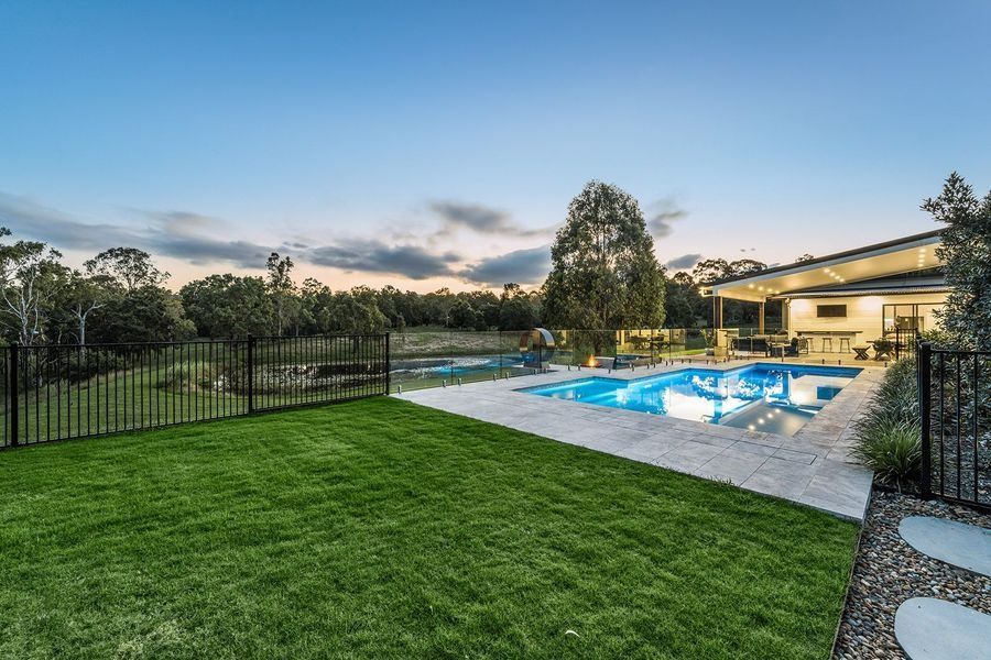 Backyard with pool, green lawn, black fence, and trees under a dusk sky.