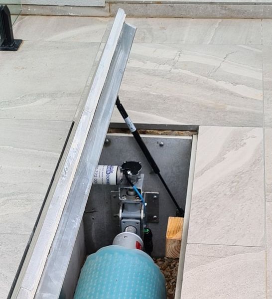 Open access panel in tiled floor, revealing machinery. A silver lid is propped open by a gas spring.