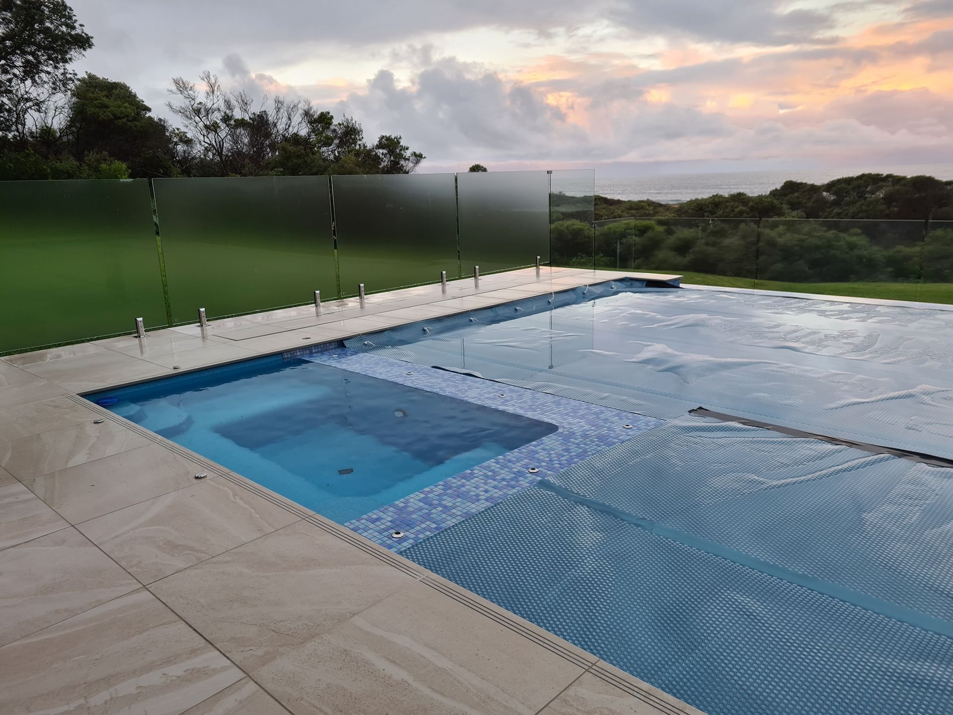 Swimming pool with glass fence, overlooking the ocean at sunset.