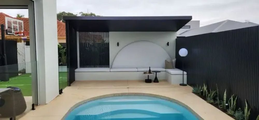 Outdoor patio with pool, black and white structure with seating, small table, and plants. Cloudy day.