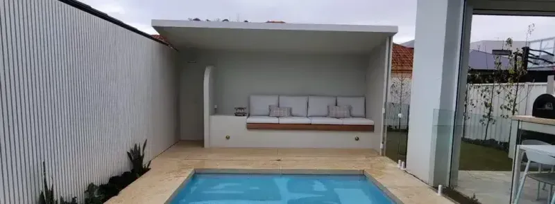 Outdoor patio with a pool, seating area under a pergola-like structure, and a white fence.