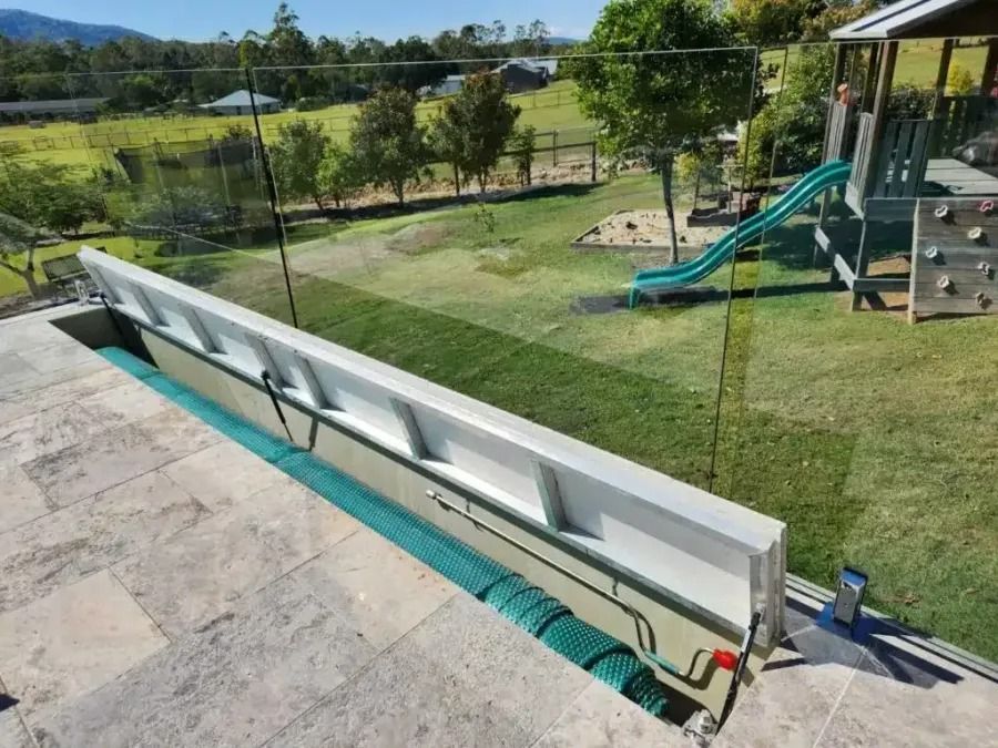 Long, light-colored barrier alongside an in-ground pool, overlooking a green yard and playground.