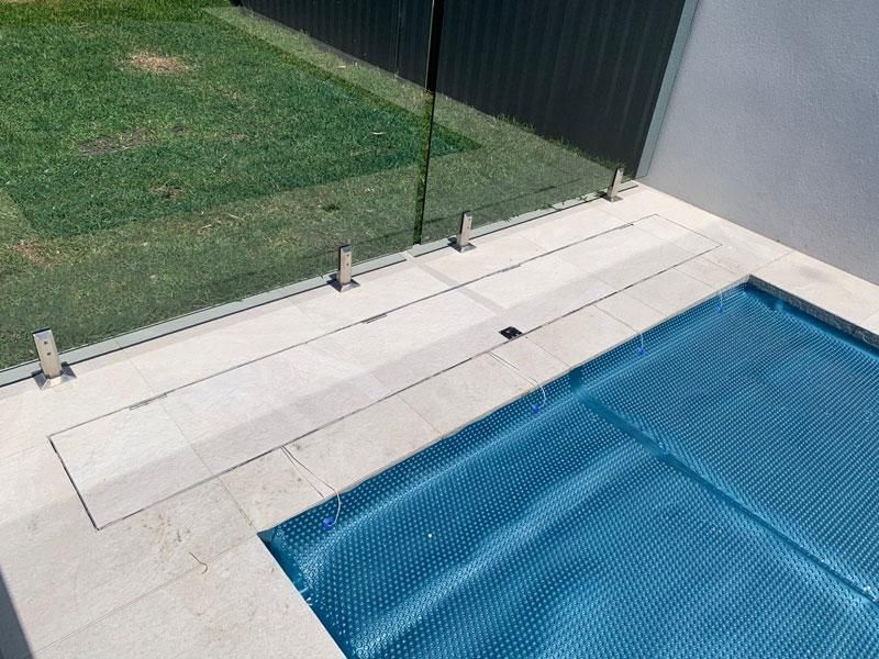 Poolside with glass fence, blue pool cover, and stone paving. Green grass and a dark fence in the background.