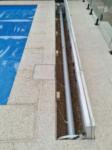 Pool cover mechanism in a narrow trench beside a pool, with a rolled-up cover visible on the pool.