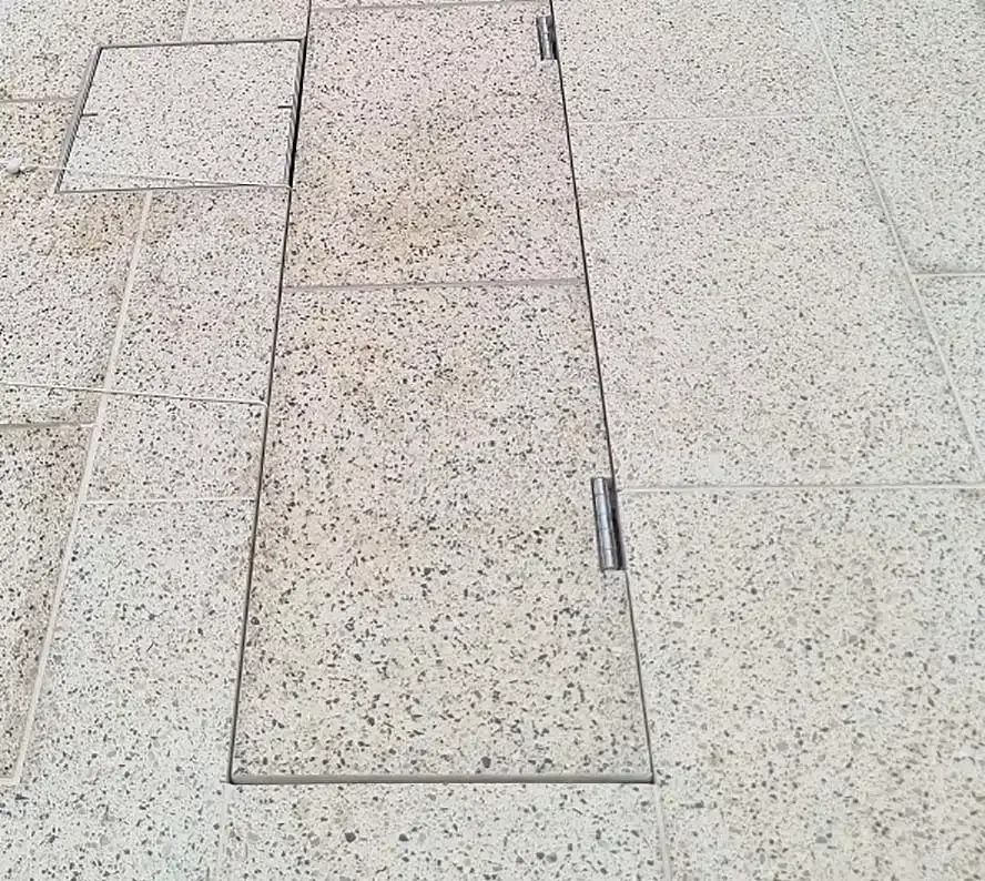 Sidewalk with speckled stone tiles, metal access covers, and hinges.