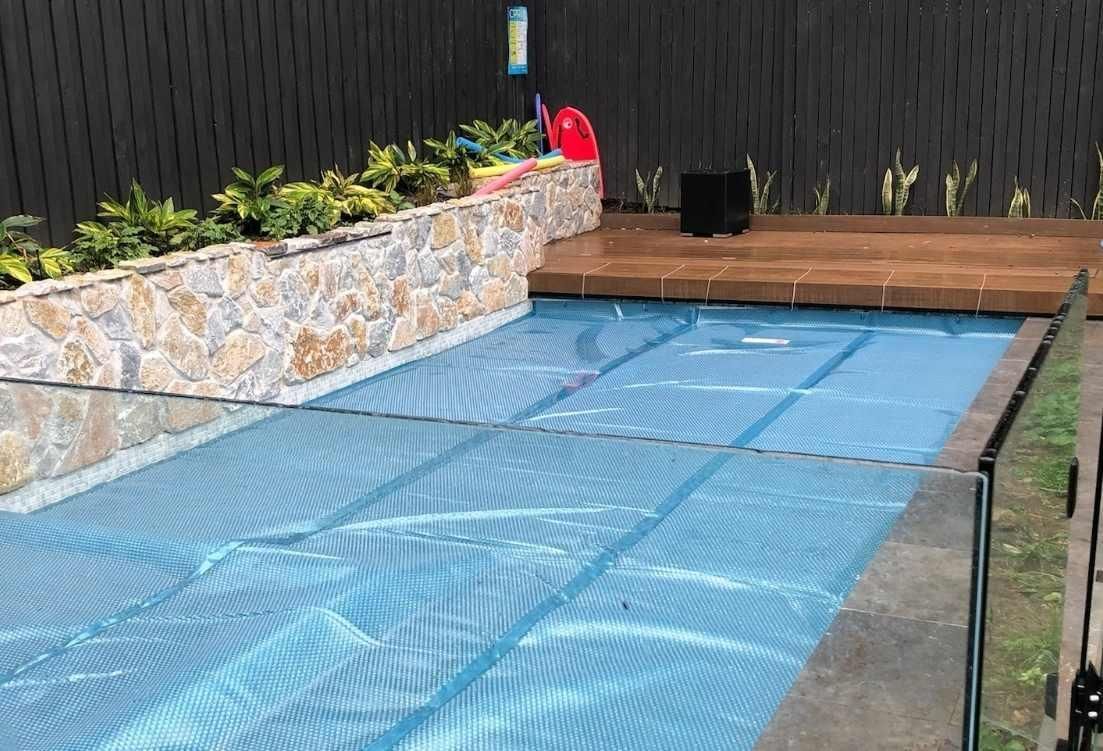 Pool covered with blue netting, surrounded by stone wall, wooden deck, and black fence.
