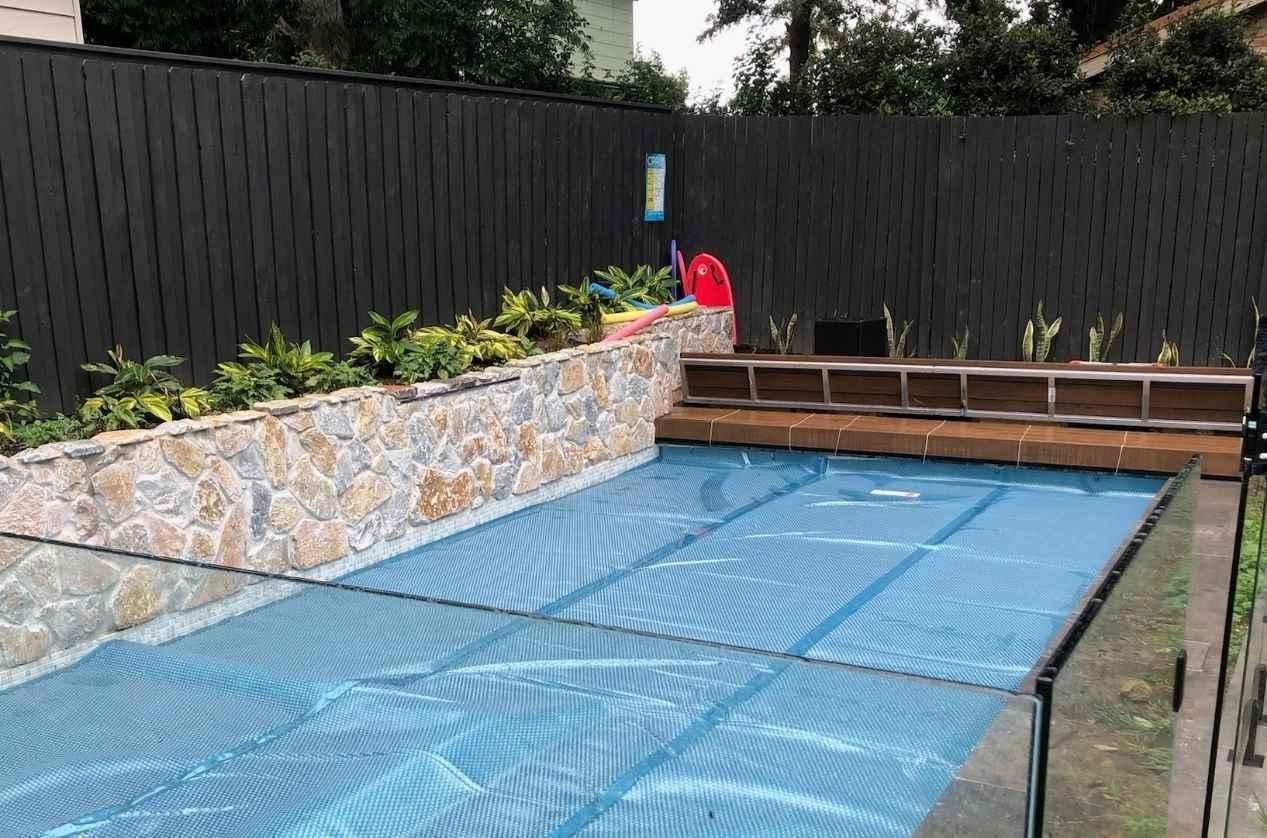 Pool with blue cover, stone wall, dark fence, and wooden deck seating.