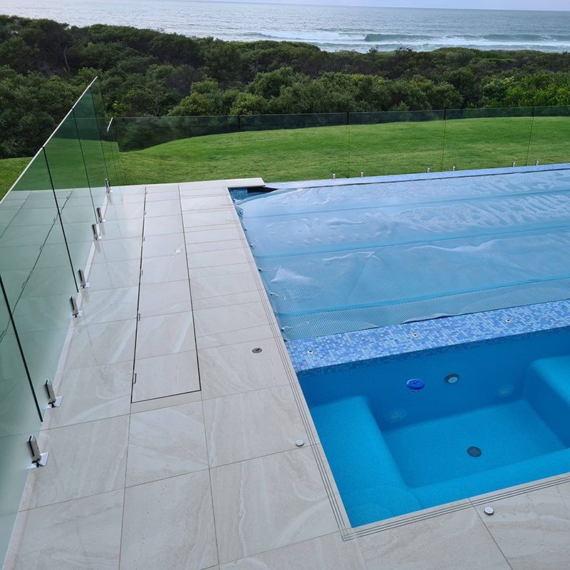 Pool with blue water and cover, glass fence, grass and ocean in background.