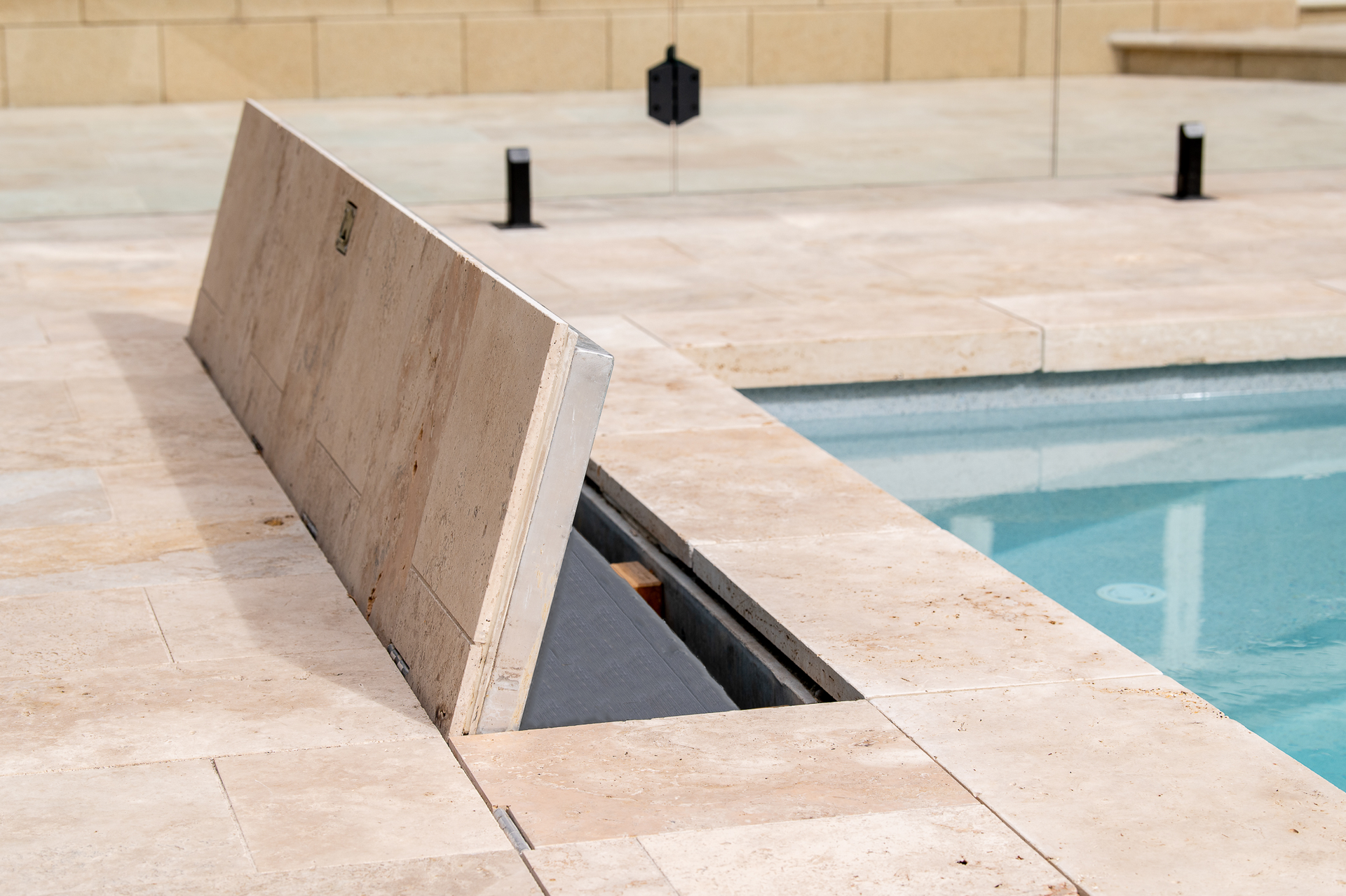 Poolside with a stone-covered automatic pool cover partially open. Light stone, water.