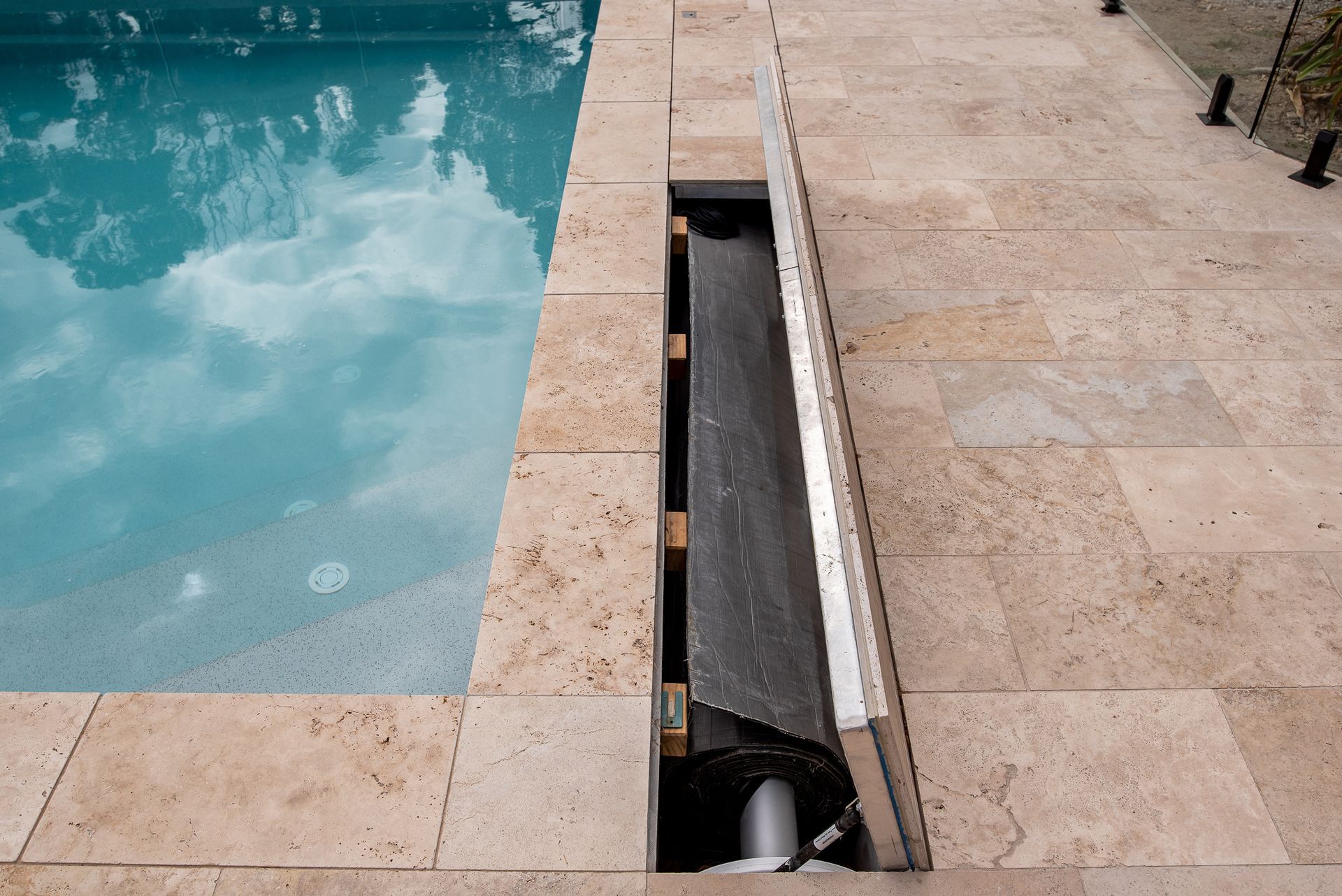 Wooden deck with a swimming pool. Gray deck with a matching wall next to the blue water. Fence in the background.
