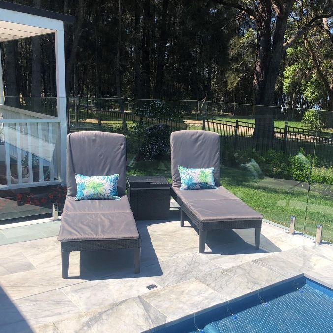 Two gray chaise lounges with blue pillows on a stone patio by a pool.