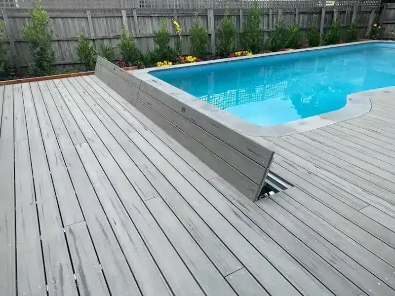Wooden deck and grey pool border near a blue swimming pool, greenery in the background.