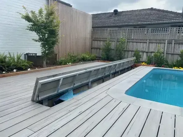 Wooden pool deck with a retractable cover revealing the pool. Landscaped backyard with trees and fencing.