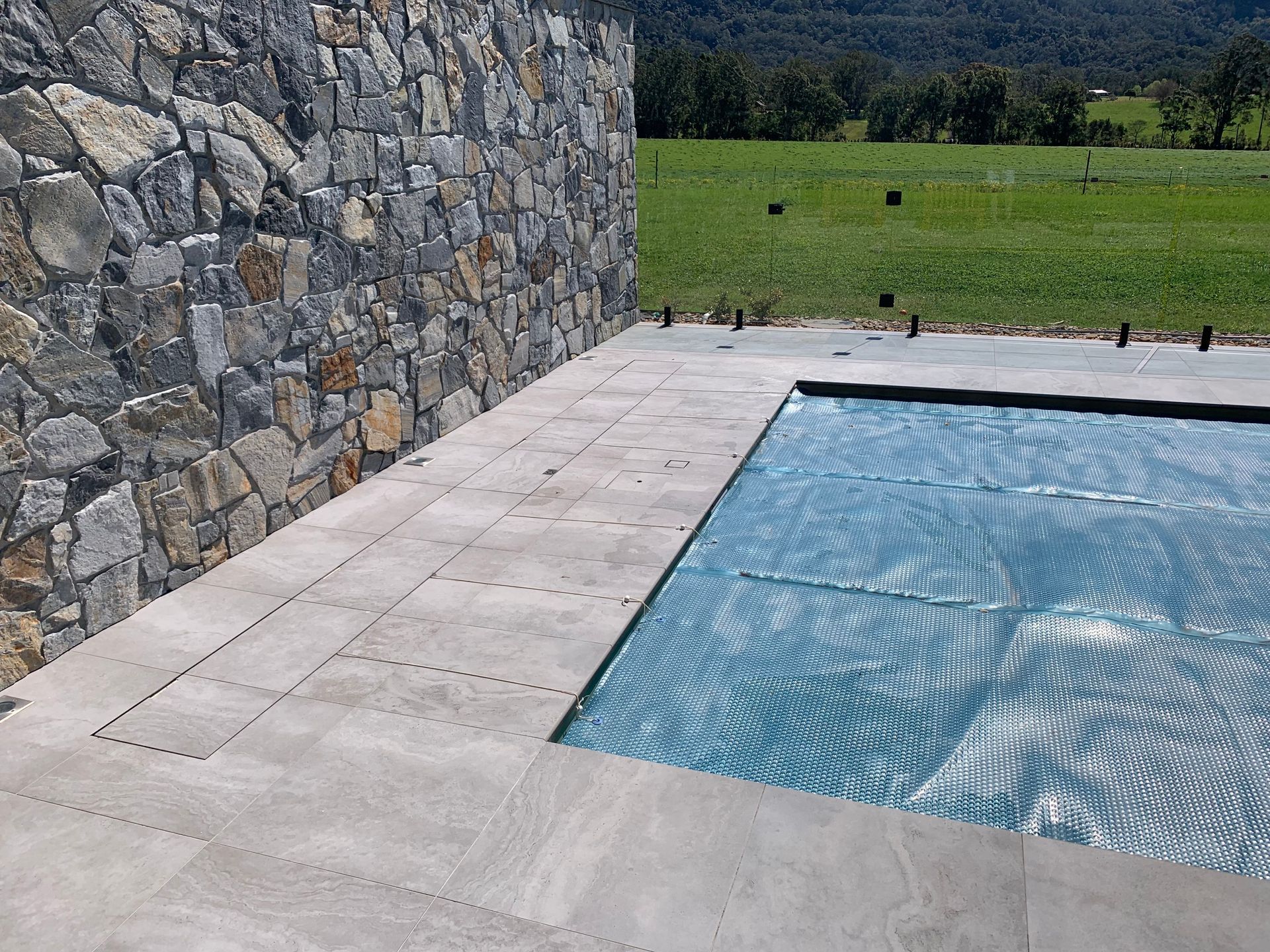 Rectangular blue swimming pool surrounded by stone tiles and a green hedge under a bright sky.