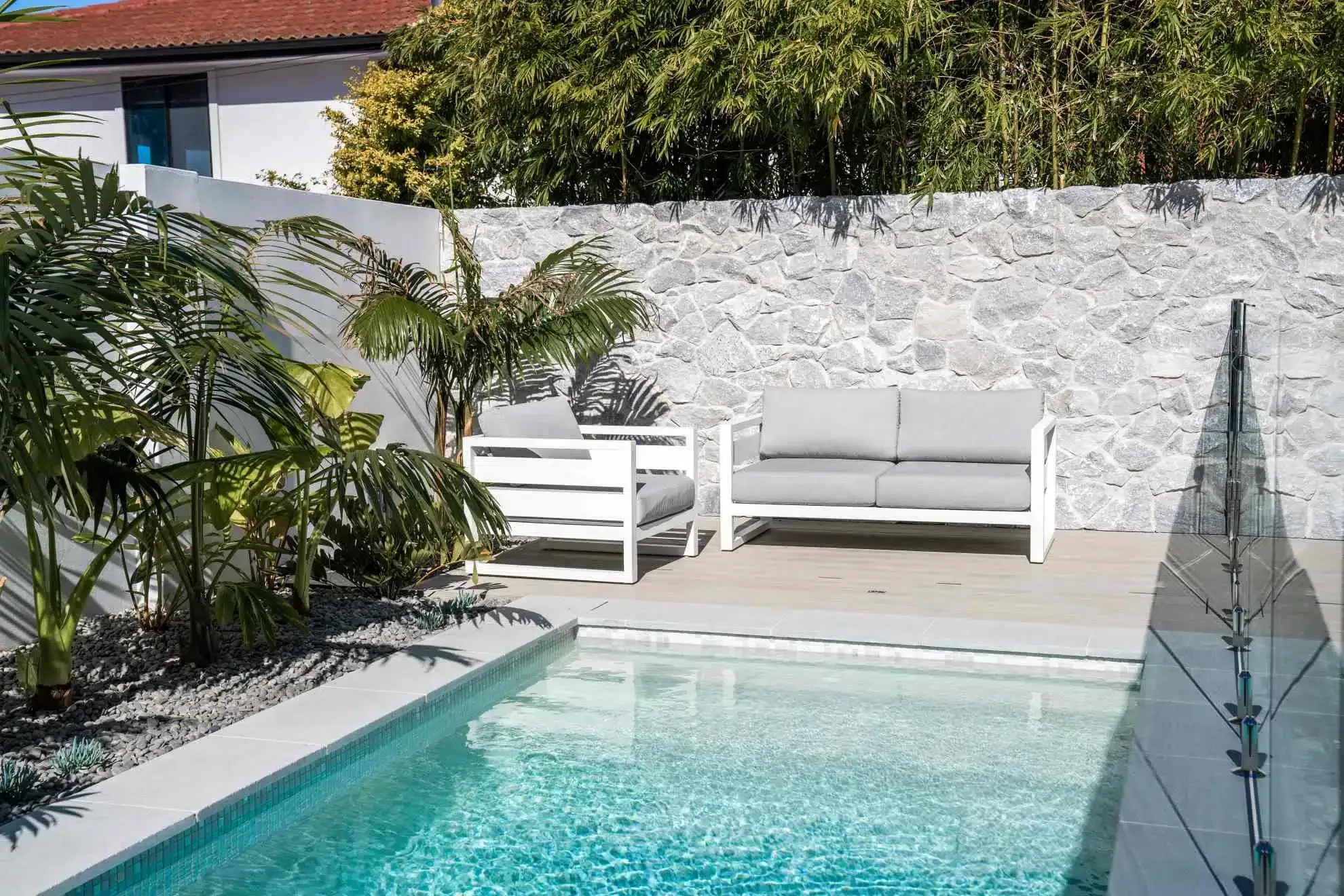 Pool with white lounge furniture on a wooden deck, a stone wall in background, greenery on either side.