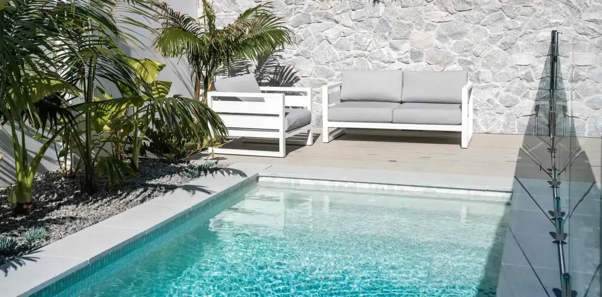 Outdoor seating area with a pool and lush greenery. White furniture sits on a wooden deck. Stone wall backdrop.