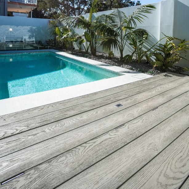 Wooden deck beside a turquoise pool, with white trim and wall, tropical plants.