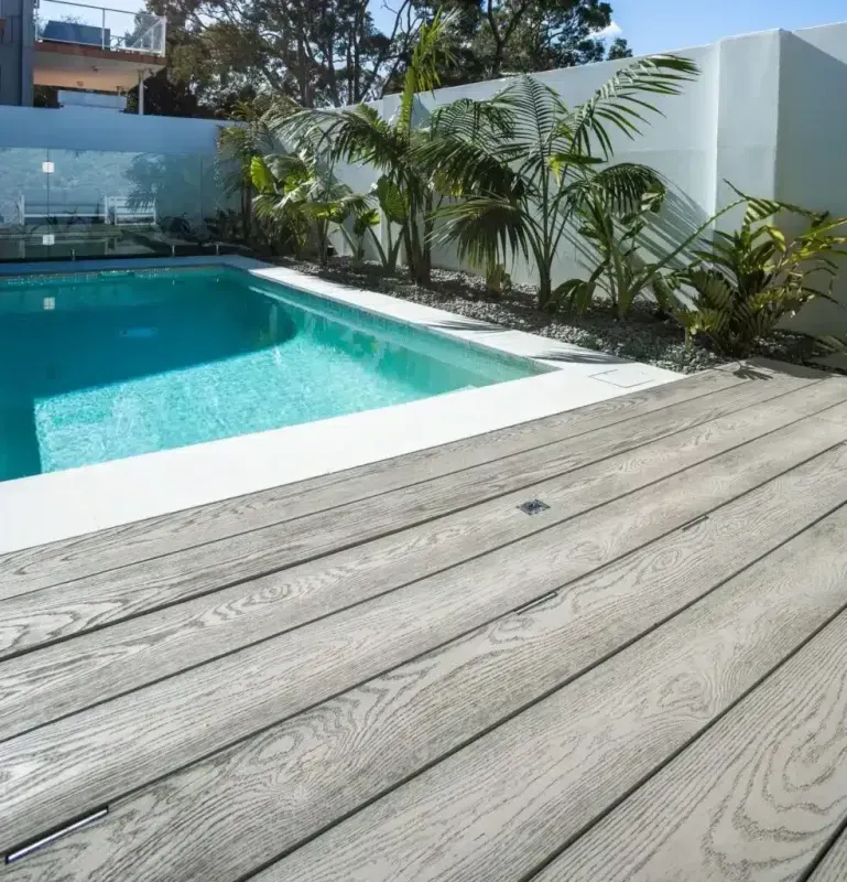 Poolside view with gray composite decking and turquoise water. White walls and tropical plants in background.