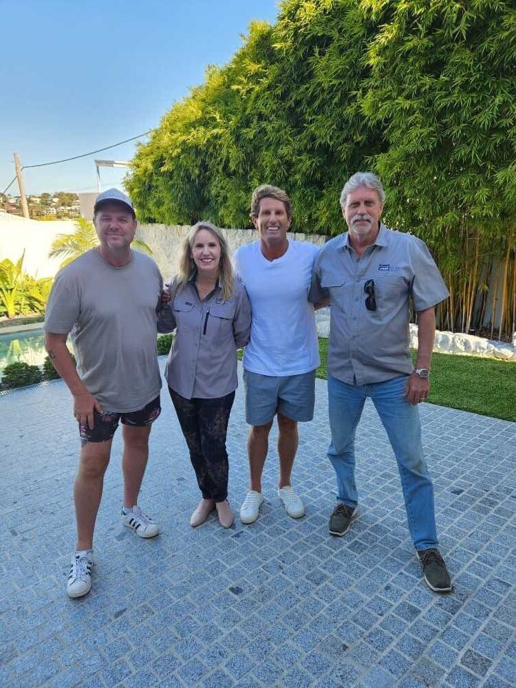Four people standing together outdoors. Two men and two women pose on stone patio near green foliage.