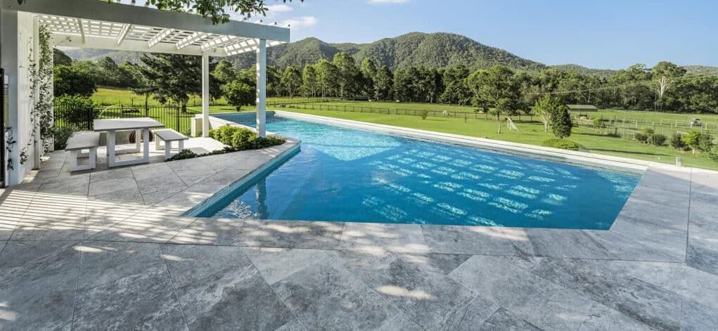 A long pool overlooks a green field and a mountain. A white pergola with a table is nearby.