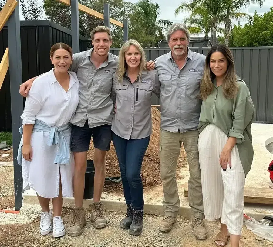 Group of five people smiling outdoors, near construction. Construction materials and a fence are visible in the background.