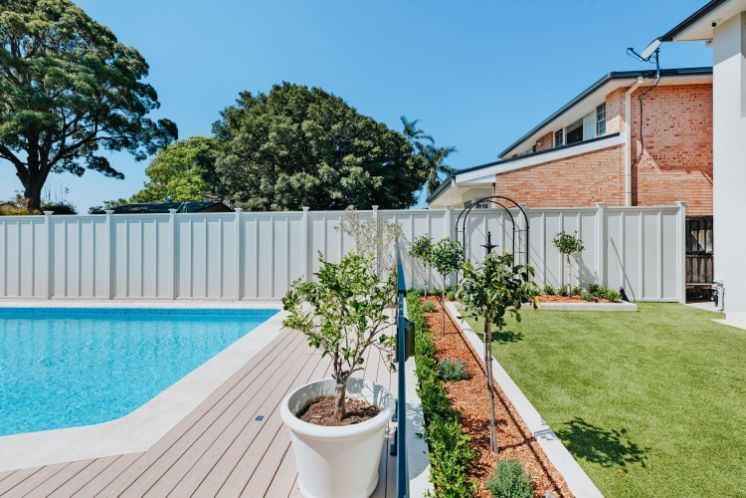 Swimming pool next to a backyard with a white fence, green grass, and trees under a blue sky.