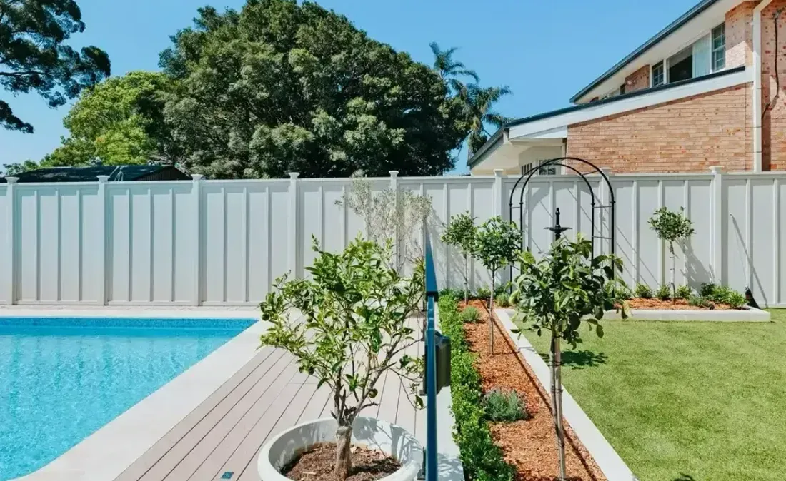 Poolside backyard with white fence, landscaping, and house in the background. Sunny day.