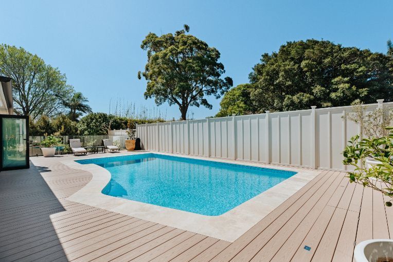 Swimming pool with surrounding wooden deck, white fence, and greenery under a blue sky.