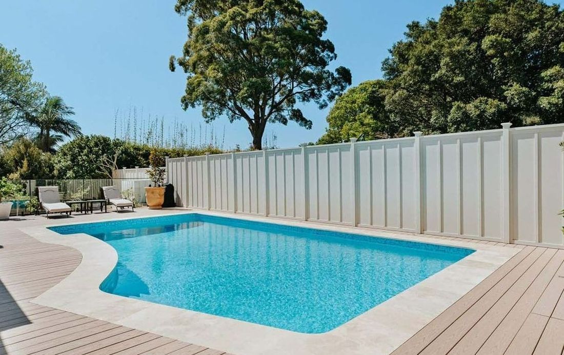 Swimming pool surrounded by a white fence, wooden deck, and lush trees under a clear blue sky.