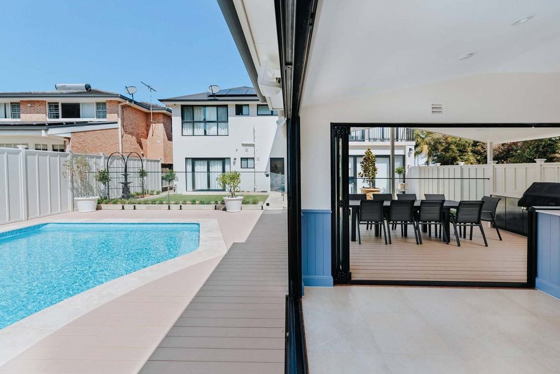 Poolside patio opens to dining area, with pool in foreground and house in the background.