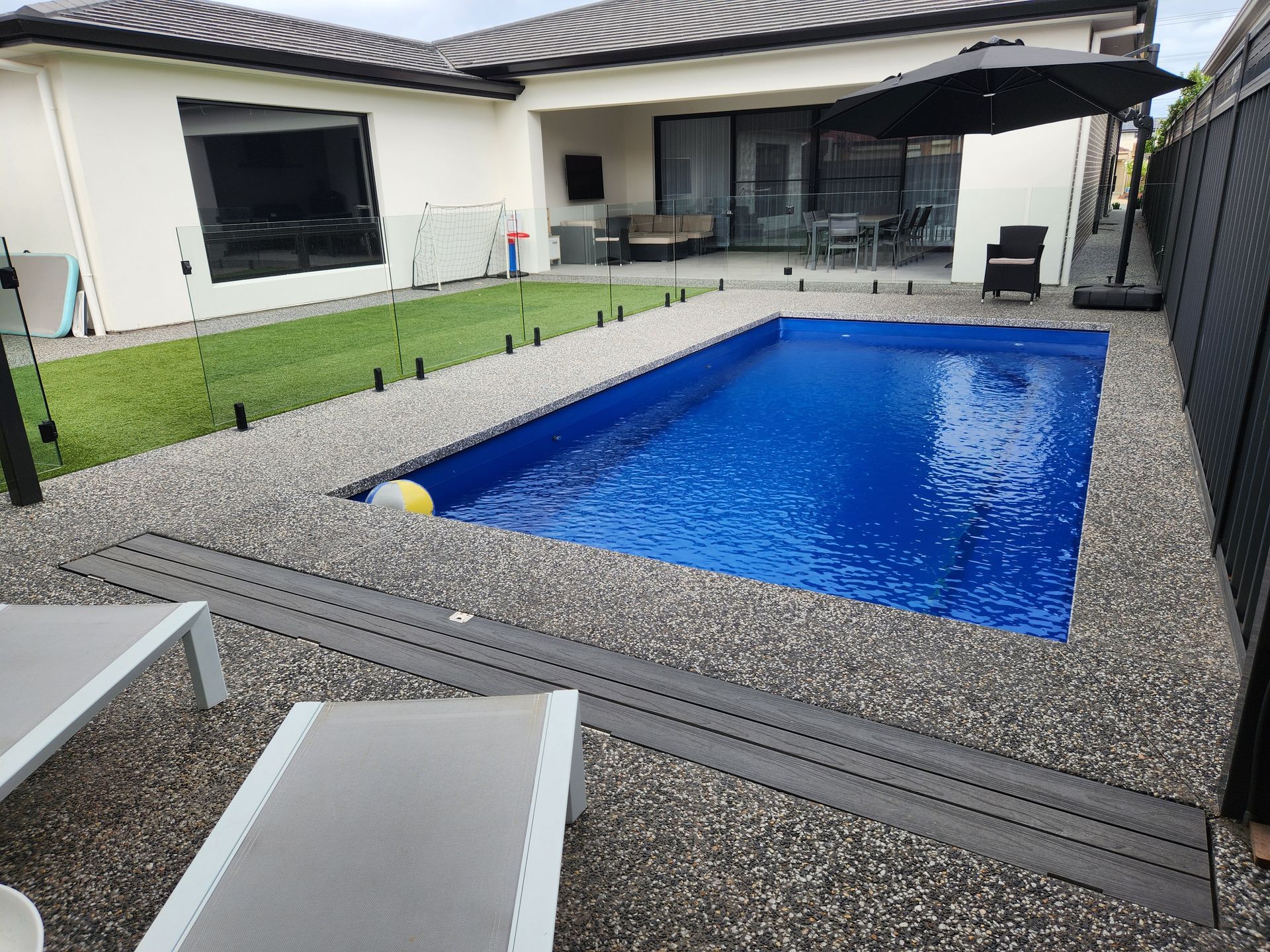Rectangular blue swimming pool surrounded by stone tiles and a green hedge under a bright sky.