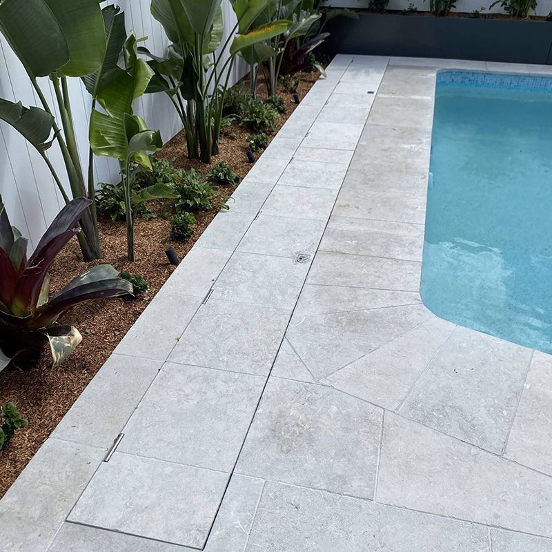 Poolside view with light-colored paving, a channel drain, garden bed with plants, and clear blue water.