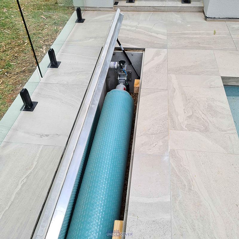 Pool cover mechanism recessed in stone decking, with rolled-up blue cover visible.