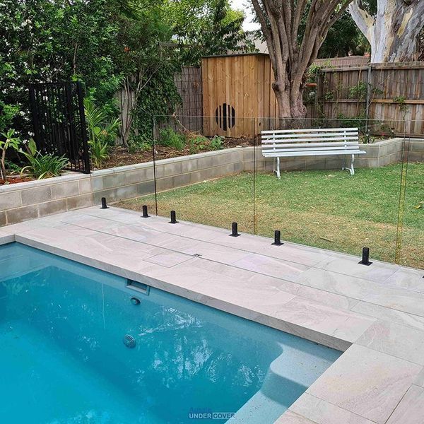 Pool with glass fence in backyard with bench and wooden structure.