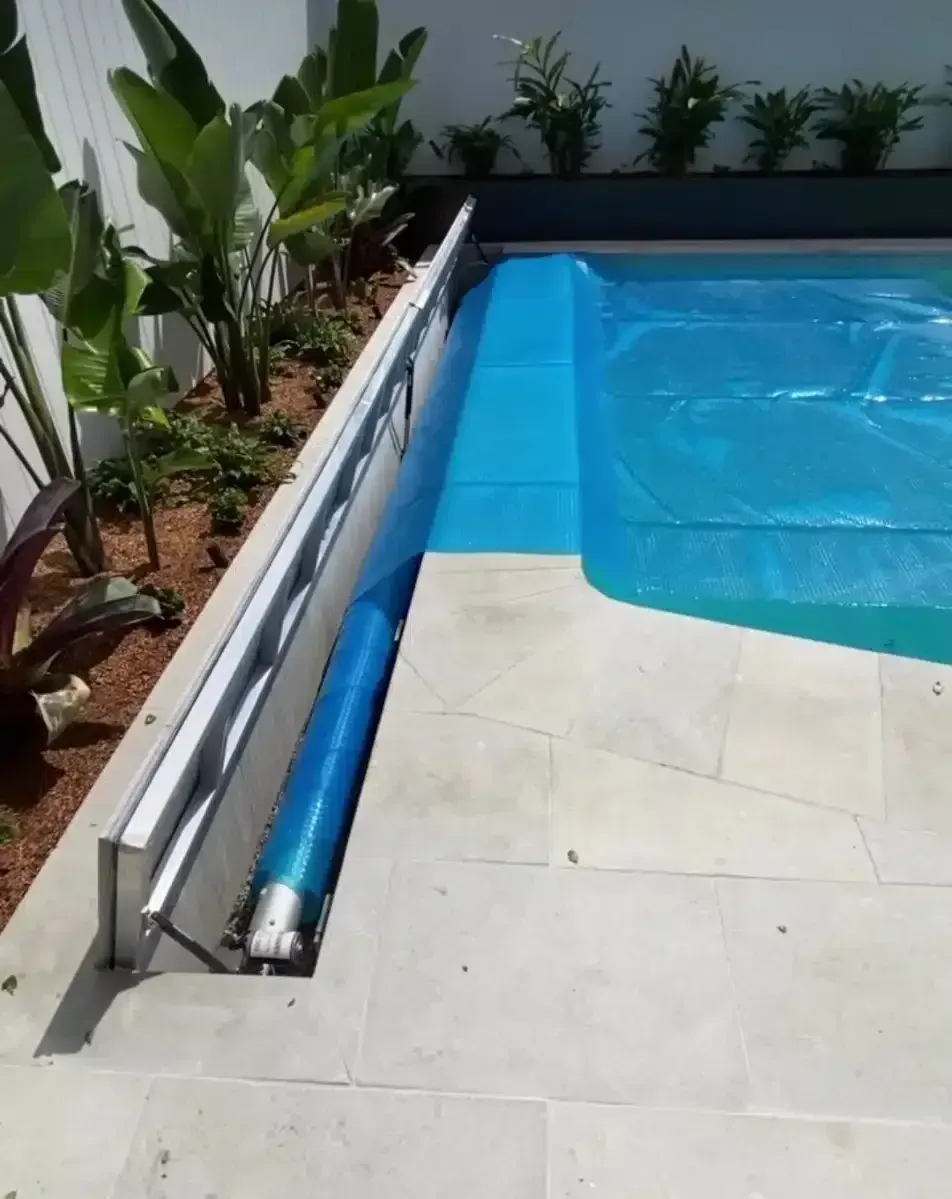 Pool cover partially open, revealing blue water. Cover rolled up in a recess, surrounded by stone and plants.