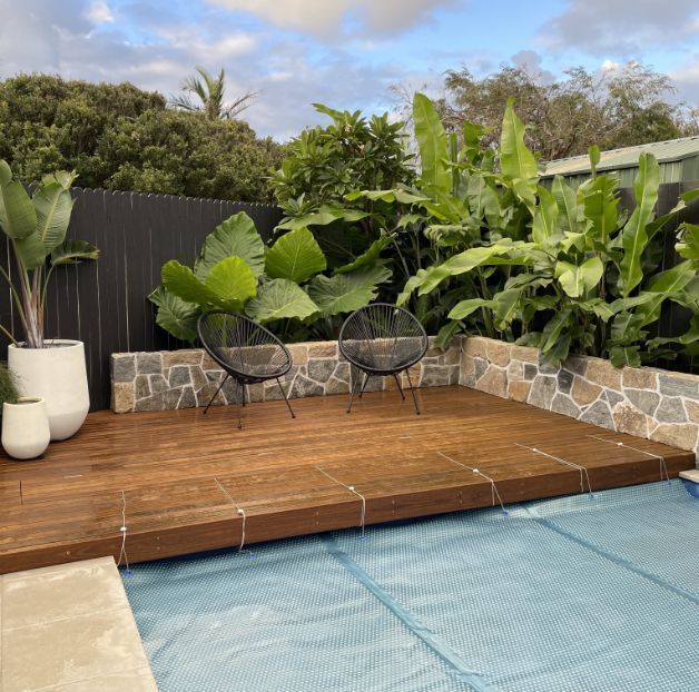 Wooden deck with two black chairs next to a pool with lush tropical plants and a stone wall backdrop.