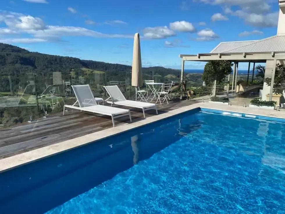 Swimming pool with lounge chairs on a deck overlooking a scenic landscape with trees, sky, and building.