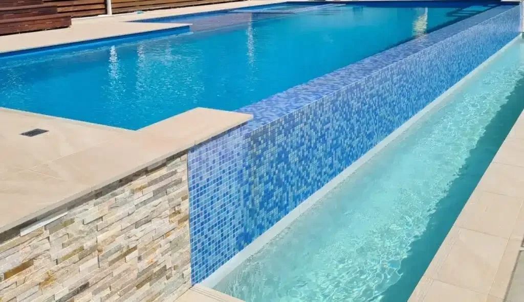 A long, rectangular swimming pool with blue tiles and clear water. Overflowing into a lower catch basin.