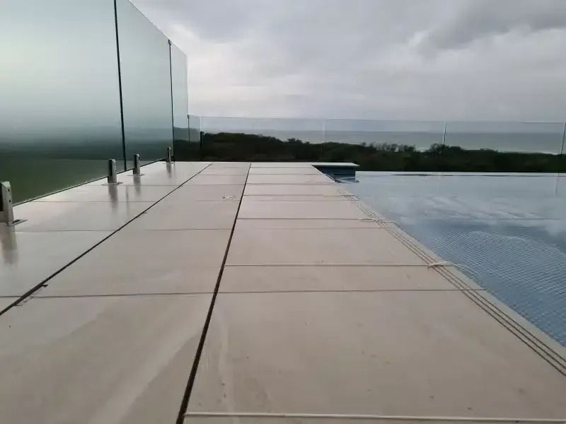 Modern pool deck with glass barrier overlooking ocean on a cloudy day.