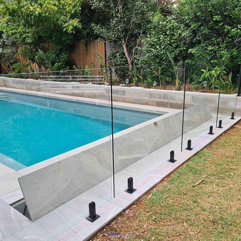 Swimming pool with glass panel fencing. Green grass, trees, and stone retaining wall.