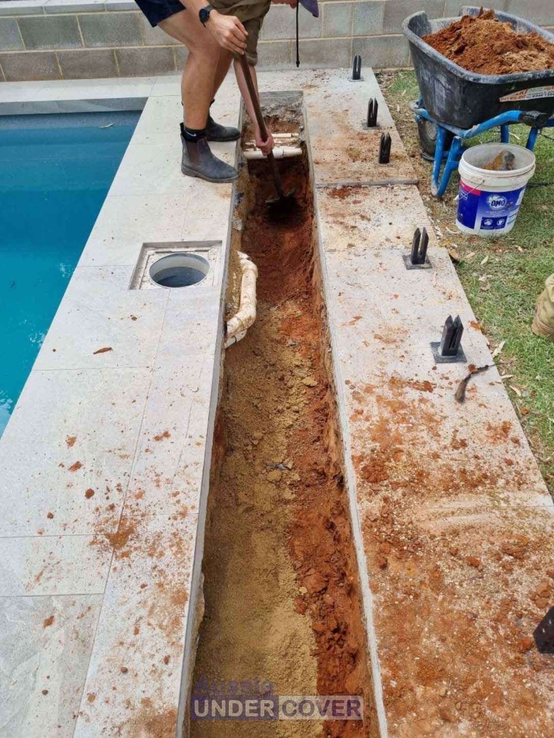 Person digging a trench next to a pool. Brown soil, stone border, and a wheelbarrow are present.